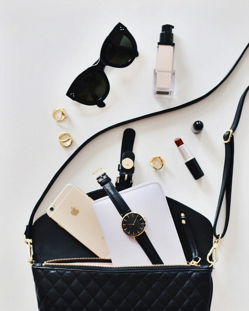 Schwarze Handtasche mit goldenen Details, darin ein Smartphone, eine Uhr und ein Notizbuch, daneben Sonnenbrille, Ringe, Foundation und Lippenstift auf weißem Hintergrund