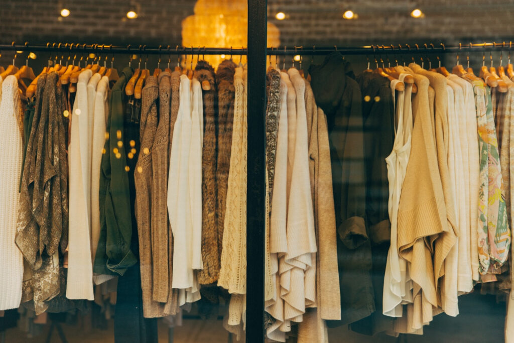 Kleiderstange mit beigen, weißen und braunen Kleidungsstücken in einem Modegeschäft, aufgenommen durch ein Schaufenster