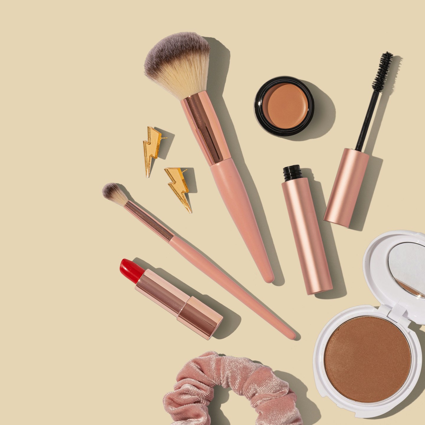 Rosa Make-up-Pinsel, roter Lippenstift, Mascara, Lidschatten, Bronzer, Haargummi und goldene Blitz-Ohrringe auf beigefarbenem Hintergrund